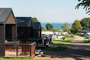 Holzhütten und Stellplätze am Campingplatz, Blick auf das Meer, Rasen und Bäume entlang des Weges.