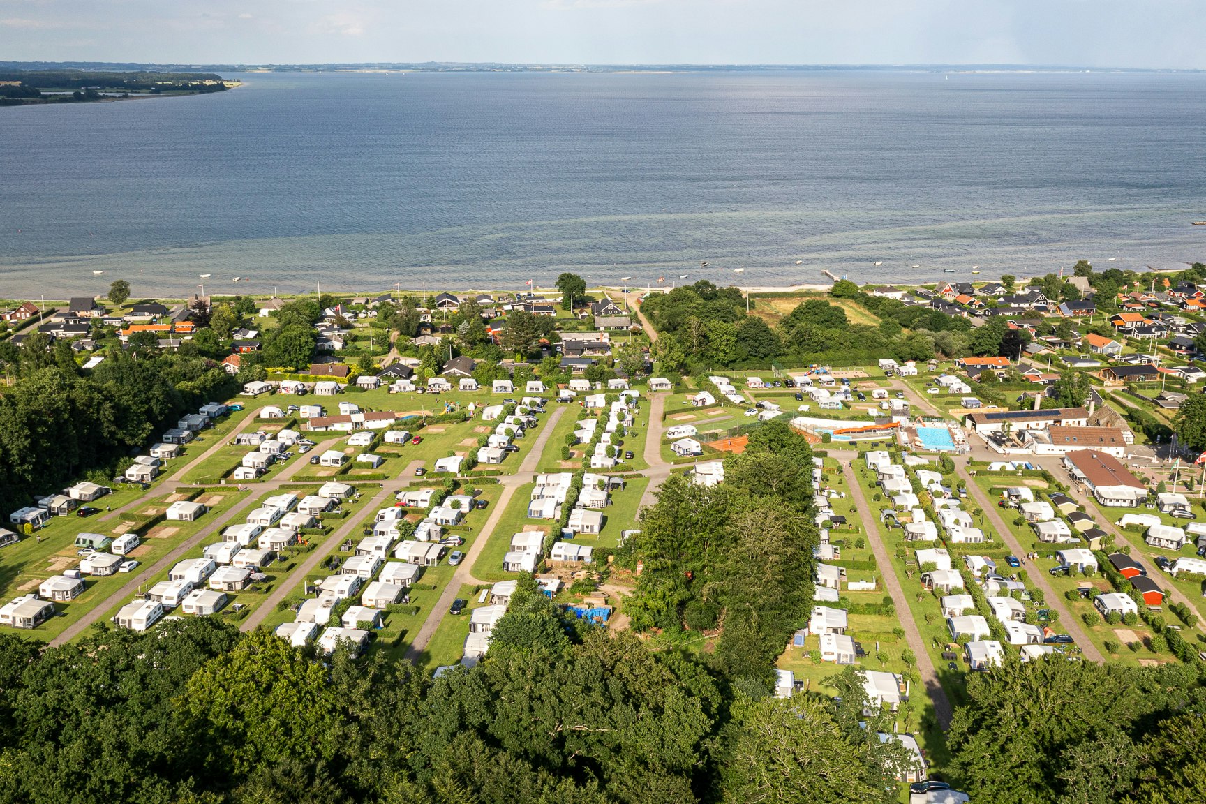 Luftaufnahme eines Campingplatzes am See mit vielen Stellplätzen, Wohnwagen und Zelten, umgeben von Bäumen und Wegen.