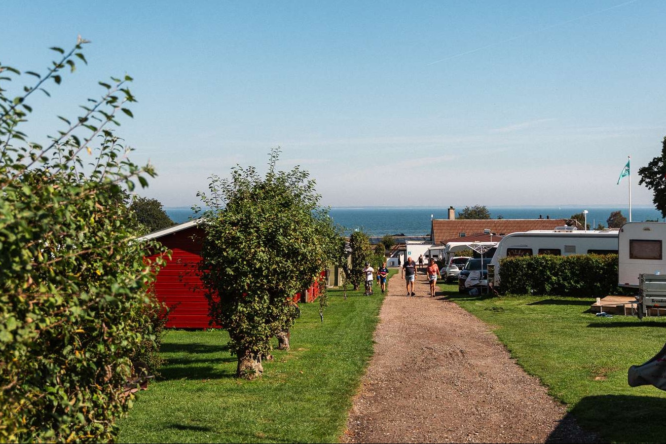 Campingplatz-Gasse mit Rasen, Hecken und Wohnwagen; im Hintergrund das Meer und ein rotes Gebäude.
