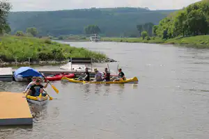 Mehrere Kanus auf dem Fluss, daneben Stege und ein Boot mit Personen, im Hintergrund bewaldete Hügel.
