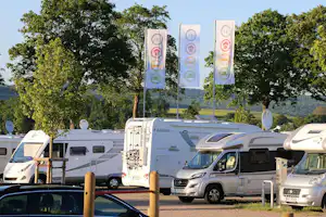 Reihe von Wohnmobilen auf einem Campingplatz, im Hintergrund Banner an hohen Masten und Bäume mit Blick auf Hügel.