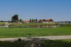 Campinganlage am Fluss mit Rasenfläche, mehreren Gebäuden mit roten Dächern, Stellplätzen und geparkten Fahrzeugen.