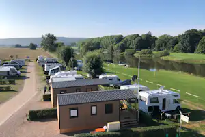 Wohnmobilstellplätze am Fluss mit Rasenfläche, Bäumen und einem Weg zwischen den Fahrzeugen auf dem Campinggelände.