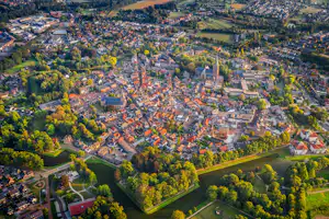 Luftaufnahme eines Dorfes mit vielen Häusern, Feldern und Wäldern, umgeben von grünen Hügeln und Straßen.