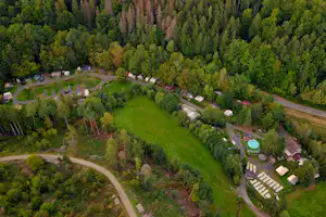 Luftaufnahme eines Campingplatzes im Wald mit Wiesen, Stellplätzen, Wegen und einem kleinen Rundbecken.