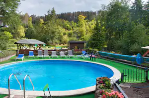 Blauer Camping-Pool mit Liegen, Sonnenschirm und Zaun, im Hintergrund Bäume und ein kleiner Pavillon.