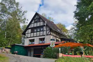Fachwerkhaus mit Satteldach, Terrasse mit Markise und Garten, daneben grüner Schuppen und Bäume im Hintergrund.