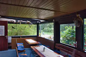Überdachte Camping-Terrasse mit Holzdach, Sitzbank und Tisch, davor Fenster und Blick auf Grün im Hintergrund.
