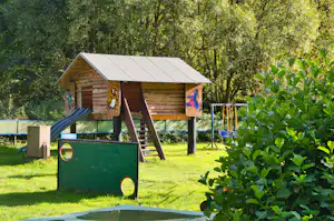 Spielhaus mit Rutsche und Holzverkleidung, im Garten mit Rasen, Bäumen und bunten Spieltafeln.