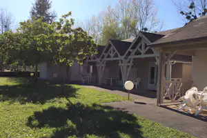 Campingunterkunft mit Veranda, weißen Stühlen und Tisch, gepflastertem Weg und Rasen im Garten mit Bäumen.