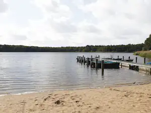 Seeufer mit Sandstrand, Steg und mehreren Bootsstegen im ruhigen Wasser, im Hintergrund bewaldete Uferlinie.