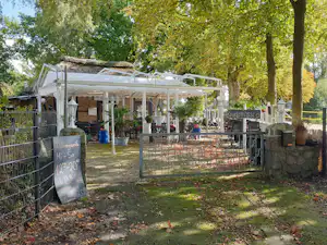 Überdachte Camping-Terrasse mit Sitzgelegenheiten, Zaun und Schildern, im Schatten von Bäumen im Außenbereich.