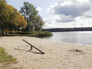 Sandstrand am See mit geländerter Seilbahn, Bäumen am Ufer und Blick auf Wasser und Wolken.