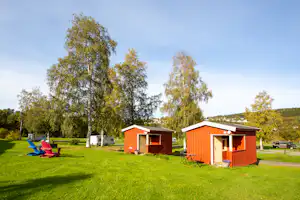 Rote Holz-Hütten auf einer grünen Wiese, davor Stühle und Tische, im Hintergrund Wohnmobile und Bäume.