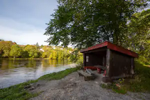 Holzhütte am Flussufer, mit Sitzbank und Steinen am Wegesrand, Bäume und grünes Ufer im Hintergrund.