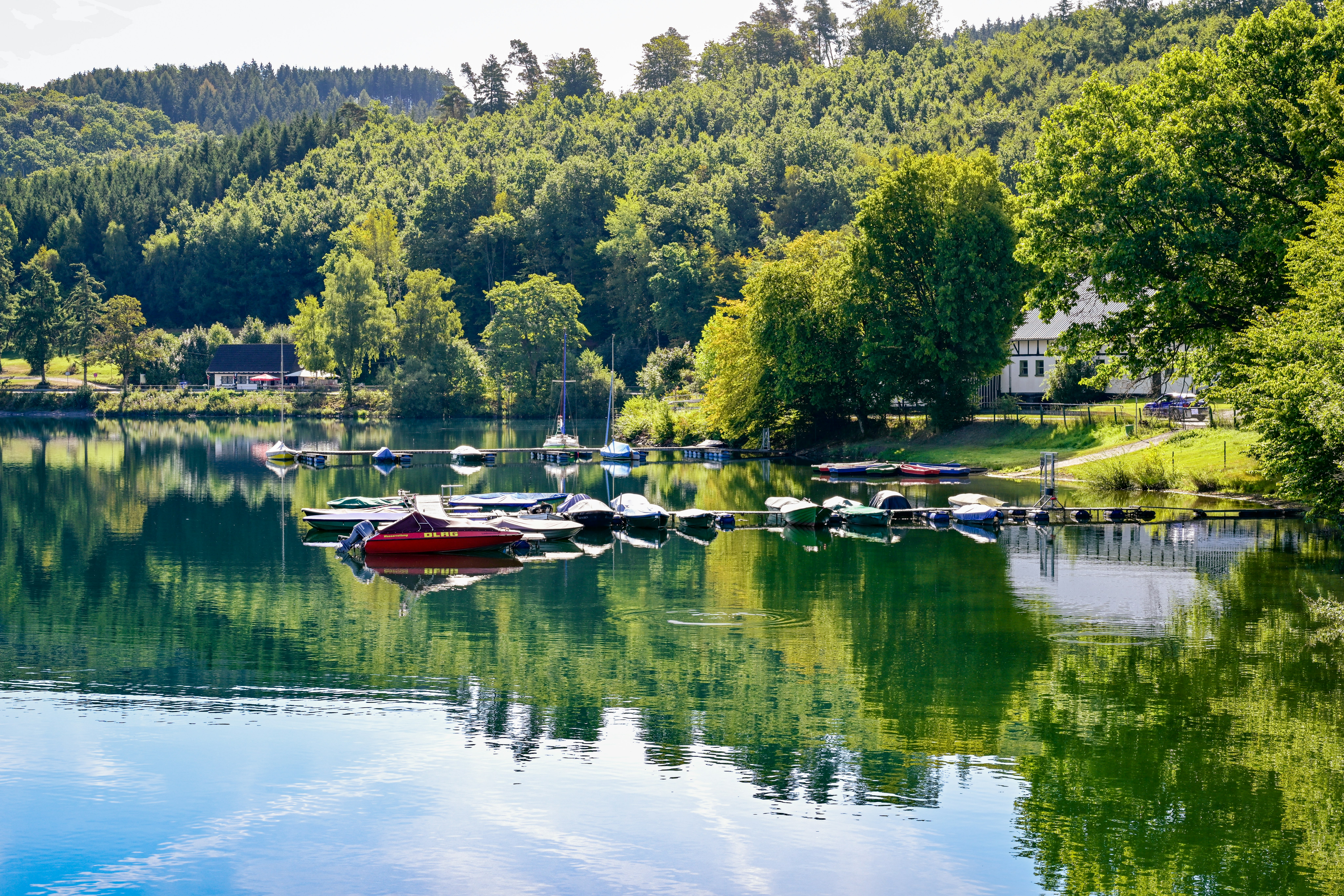 Bootssteg am See mit mehreren Booten, grünen Uferbäumen und einem kleinen Gebäude am Campingplatz.