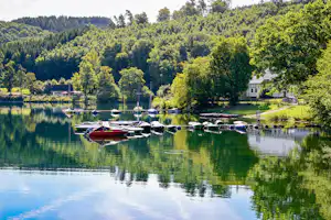 Bootssteg am See mit mehreren Booten, grünen Uferbäumen und einem kleinen Gebäude am Campingplatz.