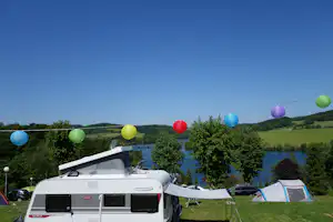 Wohnwagen und Zelte auf einem Campingplatz, im Hintergrund ein See und grüne Hügel unter blauem Himmel.