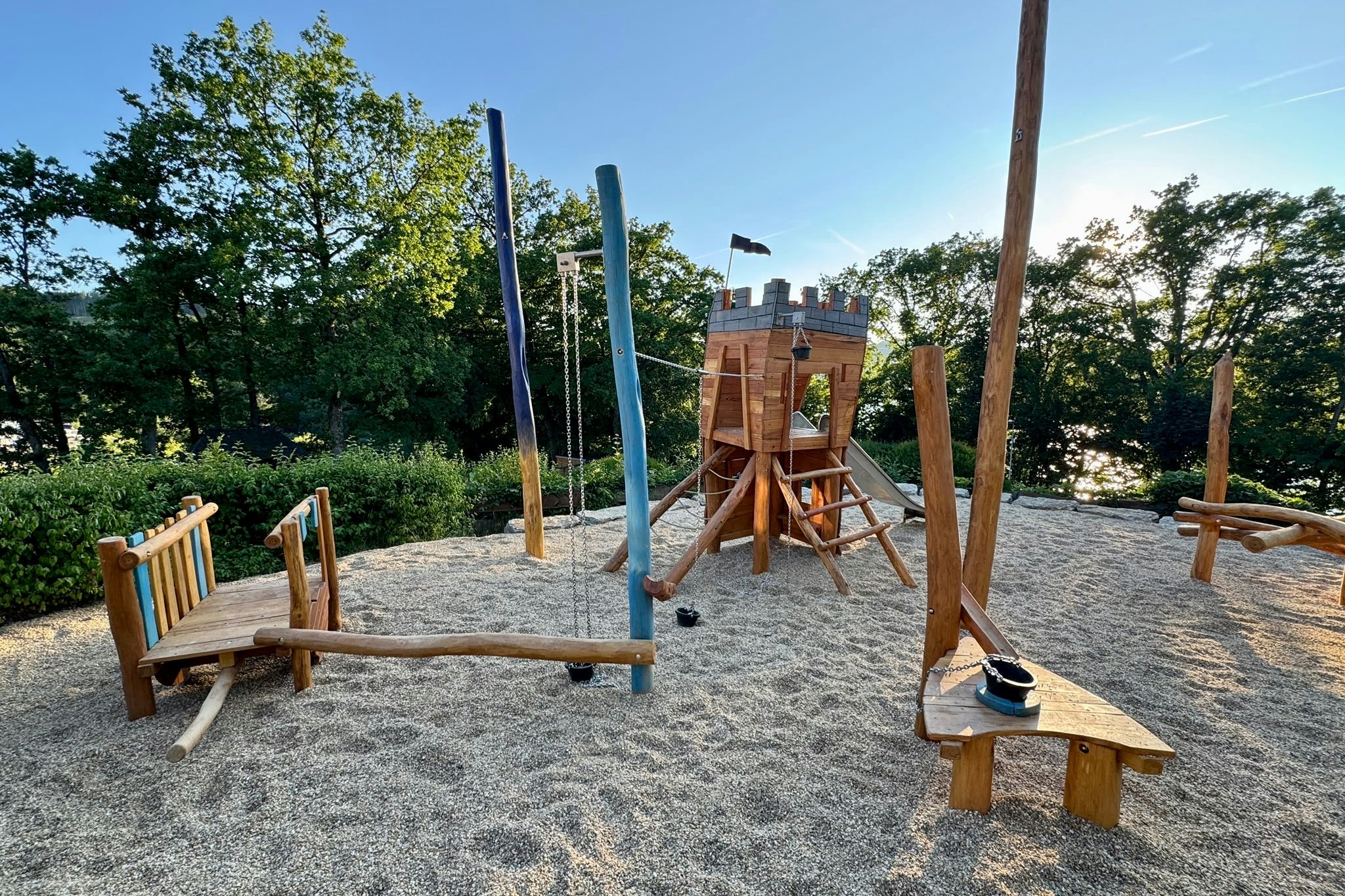 Spielplatz mit Holzschaukel und zwei Holzbänken auf einem Kiesplatz, umgeben von Bäumen und blauem Himmel.
