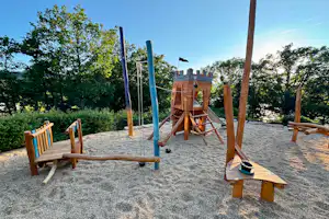 Spielplatz mit Holzschaukel und zwei Holzbänken auf einem Kiesplatz, umgeben von Bäumen und blauem Himmel.