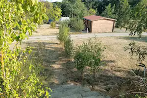 Campingplatz mit Kiesweg, Rasenfläche und kleinem Holzgebäude im Grünen, umgeben von Bäumen und Sträuchern.