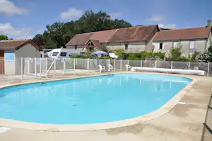 Schwimmbecken am Campingplatz mit weißem Zaun, Liegen und Blick auf Gebäude und Bäume im Hintergrund.