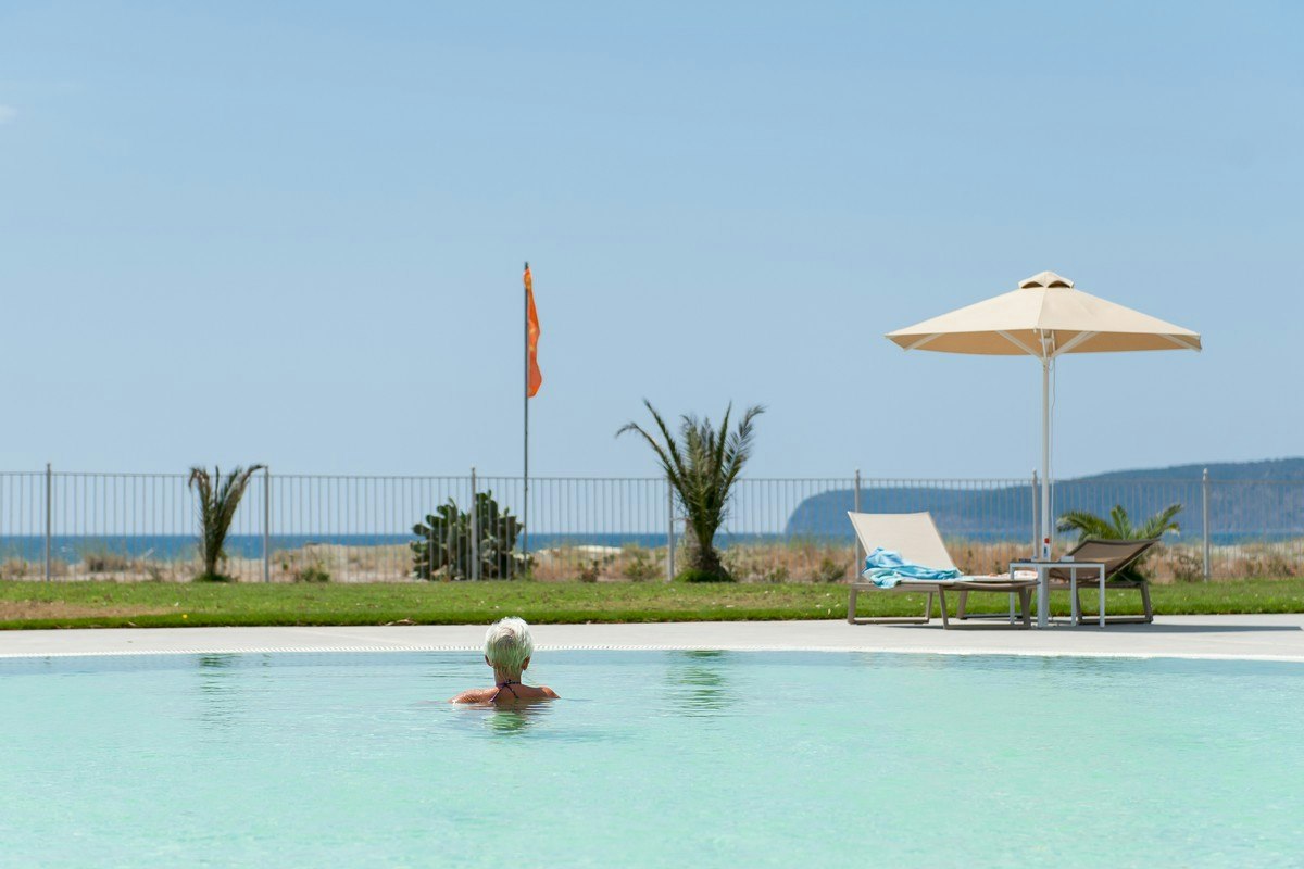Pool im Campingbereich mit Sonnenschirm und Liege, im Hintergrund Zaun, Palmen und Meerblick.