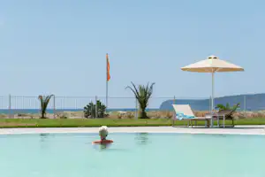 Pool im Campingbereich mit Sonnenschirm und Liege, im Hintergrund Zaun, Palmen und Meerblick.