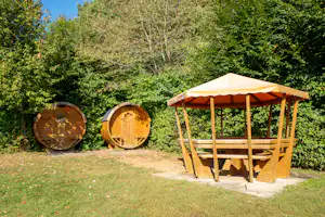 Zwei Holzfässer und ein überdachter Unterstand mit Bank und Holzstapeln im Grünen, am Wegesrand.