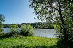 Blick auf den See mit grünen Büschen und Bäumen, im Hintergrund bewaldete Hügel und ein Schild am Weg.