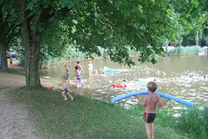 Badegäste am Seeufer mit Schwimmreifen und Luftmatratzen, im Hintergrund Bäume und ein Kiesweg am Campingplatz.