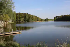 Ruhiger See mit Holzsteg am Ufer, dahinter Wald und Wiesen, im Hintergrund ein Kirchturm.