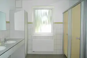 Helles Badezimmer mit Waschbecken, Fliesenwand, Fenster mit Vorhang und Heizkörper unter dem Fenster.