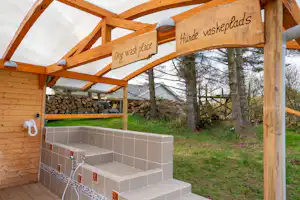 Überdachter Waschplatz mit Holzrahmen, Schildern „Dog wash“ und „Honde waskeplads“ sowie Waschbecken im Campingbereich.
