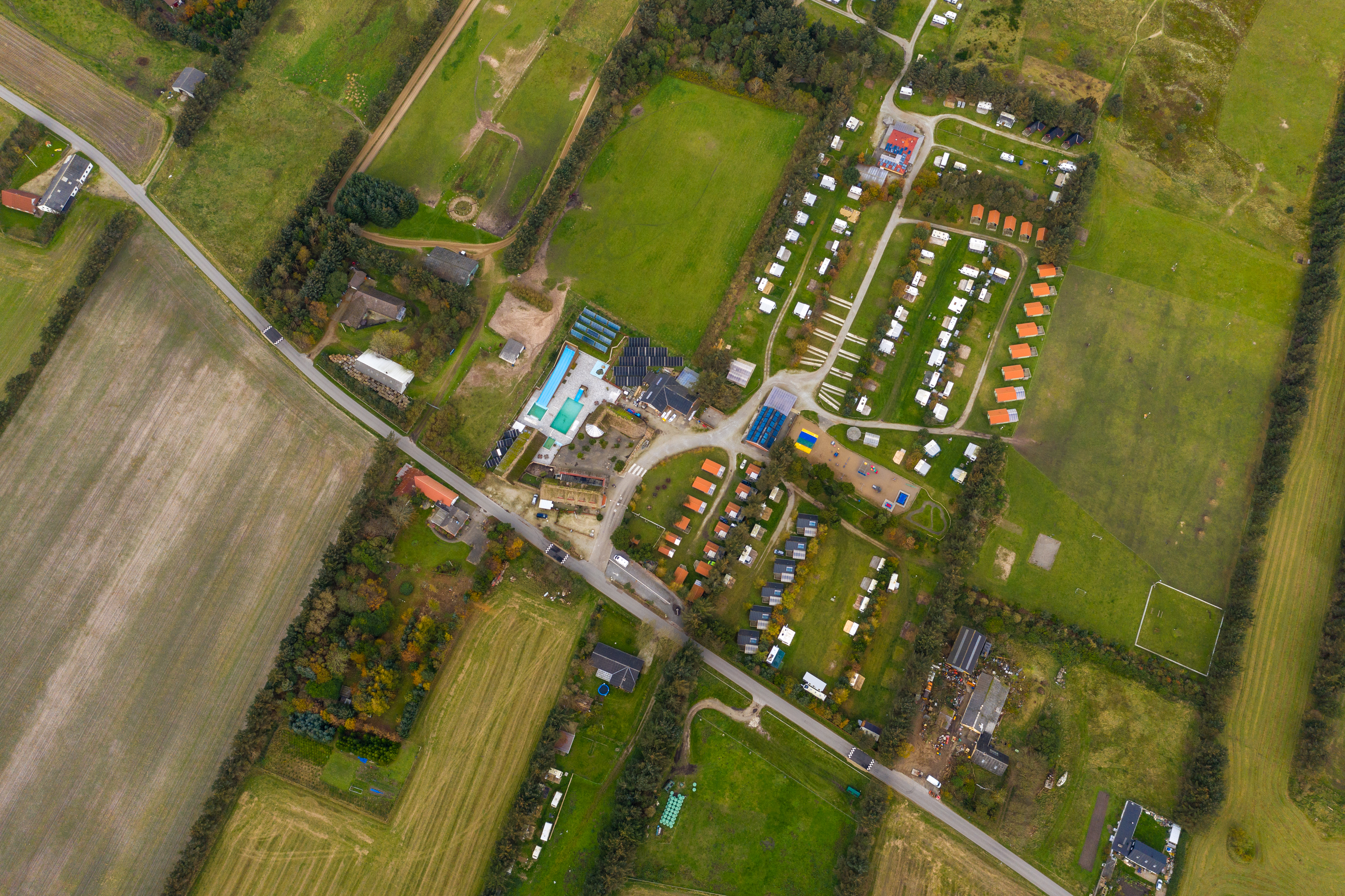 Luftaufnahme eines Campingplatzes mit Stellplätzen, Wegen und mehreren Gebäuden inmitten von Wiesen und Feldern.