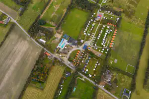 Luftaufnahme eines Campingplatzes mit Stellplätzen, Wegen und mehreren Gebäuden inmitten von Wiesen und Feldern.