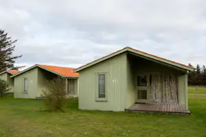 Grünes Holzhaus mit Veranda und Eingangstür, daneben ein weiteres Ferienhaus, Rasen und Bäume im Hintergrund.