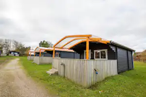 Stacaravan mit Veranda und Holzverkleidung, daneben ein Weg und Rasenfläche, im Hintergrund weitere Unterkünfte und Bäume.