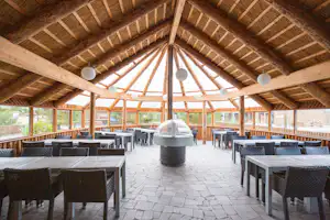 Überdachte Grillhütte mit Sitzplätzen aus dunklem Geflecht, Steinboden und Grill in der Mitte unter Holzdecke.