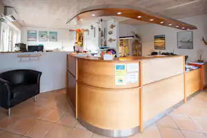 Rezeptionstresen im Campinggebäude mit Holzfront, Schreibtisch, Infomaterial und Fliesenboden, im Hintergrund Sitzbereich und Wandbilder.