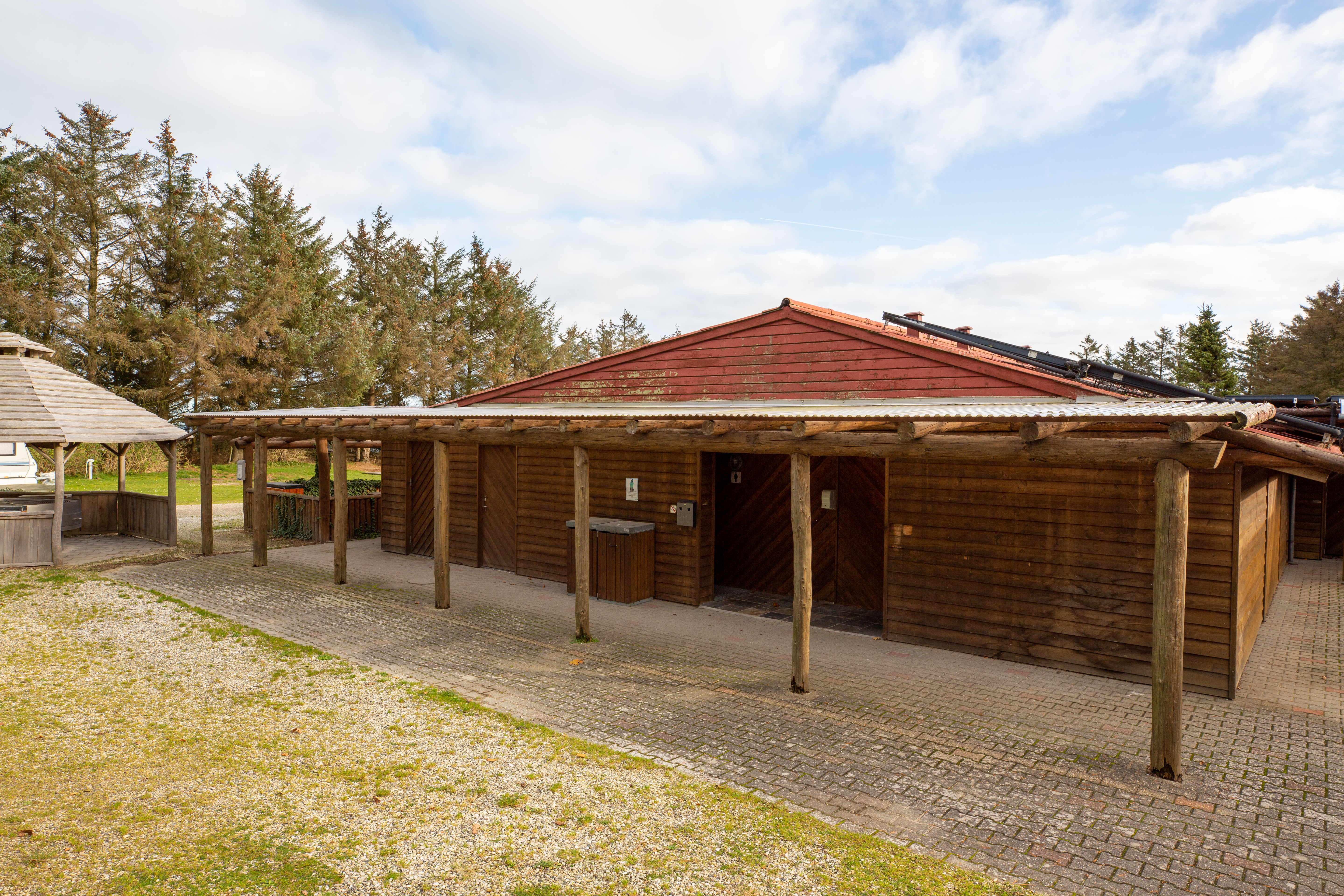 Holz-Carport am Campingplatz mit überdachter Terrasse, gepflastertem Boden und Blick auf Bäume im Hintergrund.