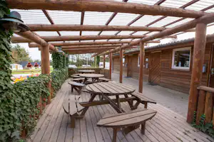 Holzterrasse mit mehreren Tischen und Bänken unter einer Pergola, daneben ein Holzgebäude am Campingplatz.