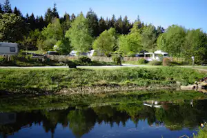 Campingplatz mit Wohnwagen und Autos auf Stellplätzen, umgeben von Bäumen, Wiese und einem kleinen Teich im Vordergrund.
