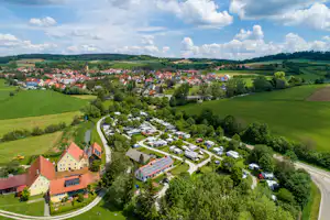 Luftaufnahme eines Campingplatzes mit Wohnwagen und Zelten, umgeben von grünen Wiesen, Feldern und einem Dorf im Hintergrund.
