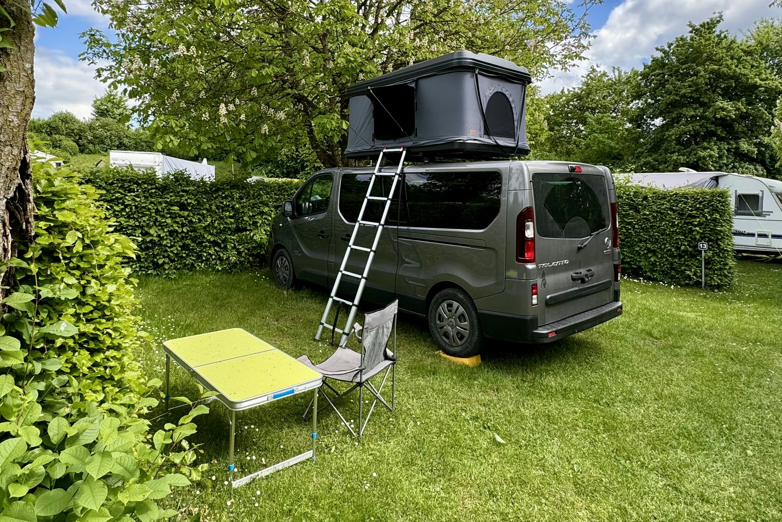 Wohnmobil mit Dachzelt auf dem Stellplatz, daneben Klapptisch und Campingstuhl im Grünen zwischen Hecken und Bäumen.