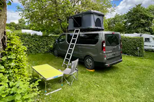 Wohnmobil mit Dachzelt auf dem Stellplatz, daneben Klapptisch und Campingstuhl im Grünen zwischen Hecken und Bäumen.
