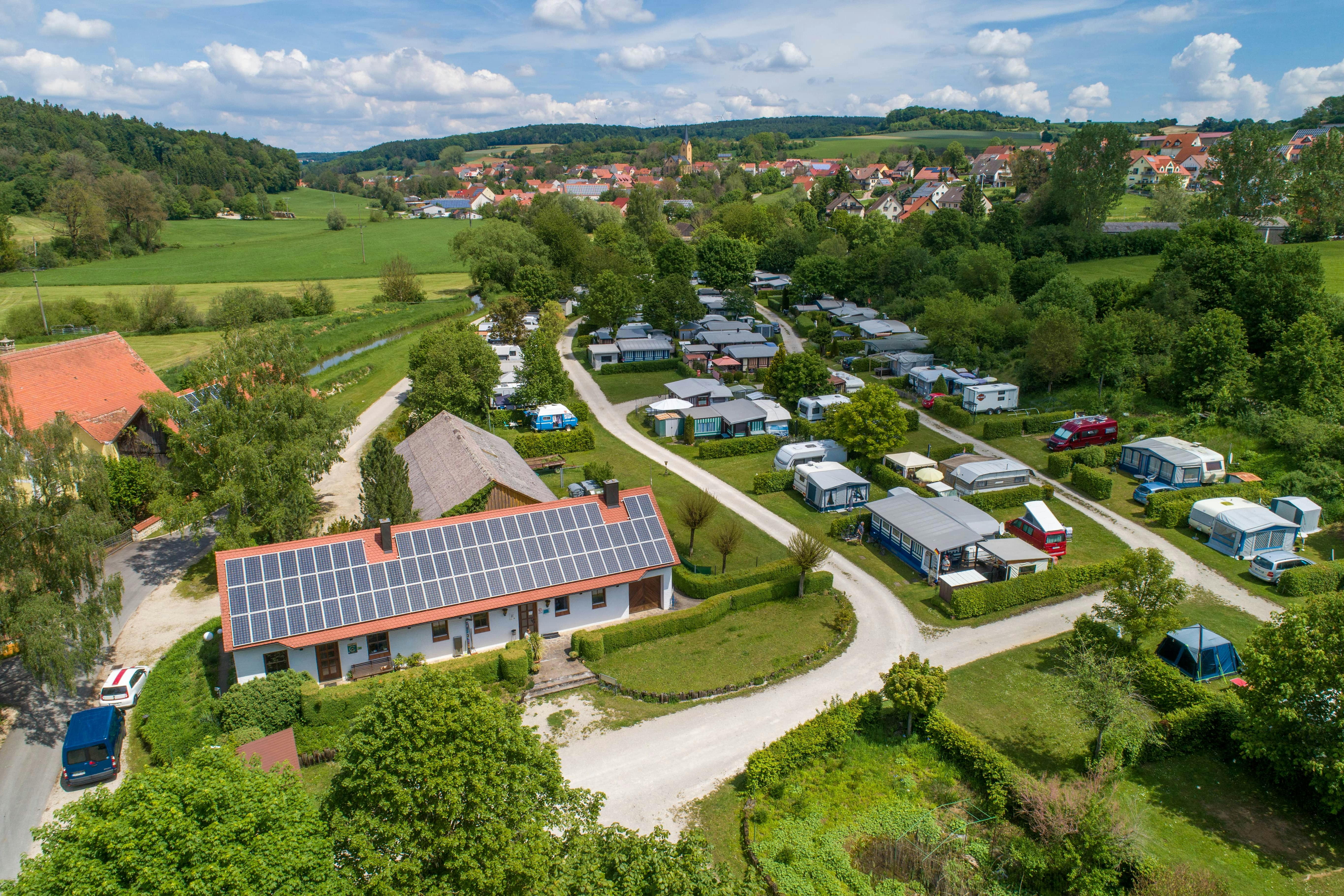 Luftaufnahme eines Campingplatzes mit Wohnmobilen und Zelten entlang der Wege, umgeben von grünen Wiesen und einem Dorf im Hintergrund.