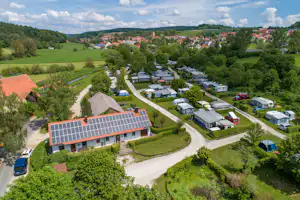 Luftaufnahme eines Campingplatzes mit Wohnmobilen und Zelten entlang der Wege, umgeben von grünen Wiesen und einem Dorf im Hintergrund.