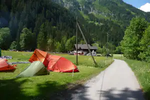 Zelte auf einer grünen Wiese am Campingplatz, daneben ein Kiesweg und Bäume vor den grünen Bergen.
