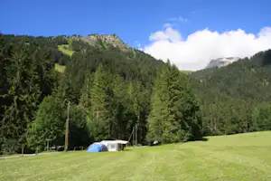 Campingzelt auf einer grünen Wiese am Waldrand, im Hintergrund Berge und bewaldete Hänge unter blauem Himmel.